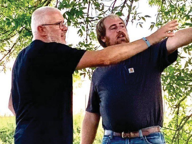 Two men standing outdoors under a tree, both wearing dark t-shirts, looking and pointing in the same direction.
